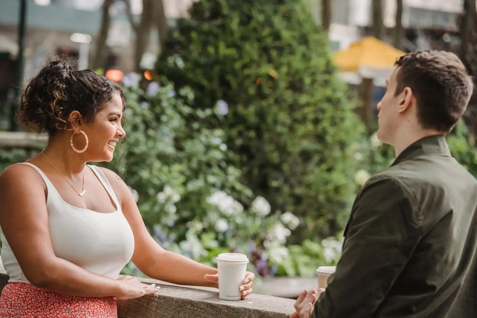 Jeune couple souriant en pleine conversation complice en extérieur, un gobelet de café à la main