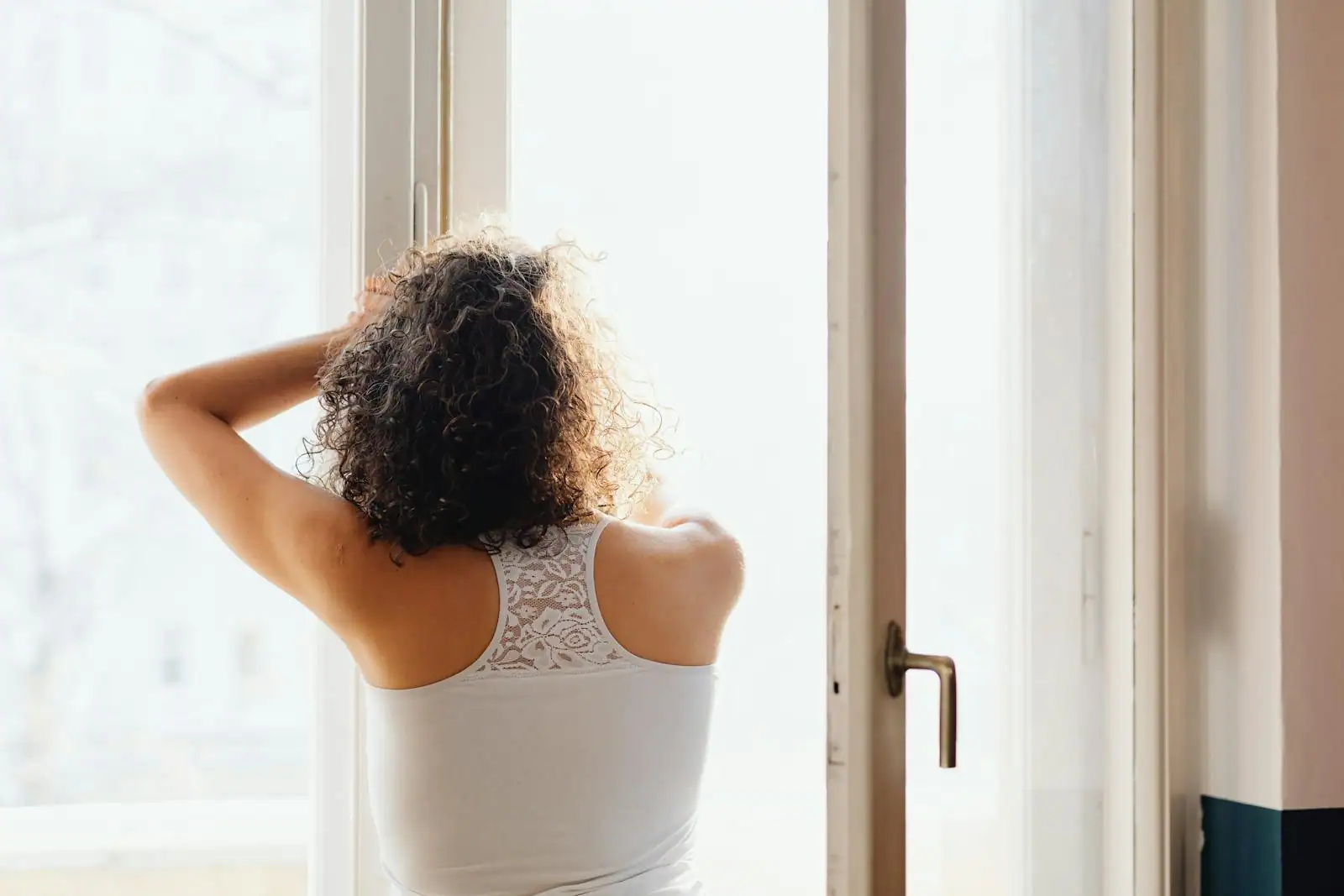 Femme debout de dos près d'une fenêtre baignée d'une lumière matinale douce, dans une posture paisible