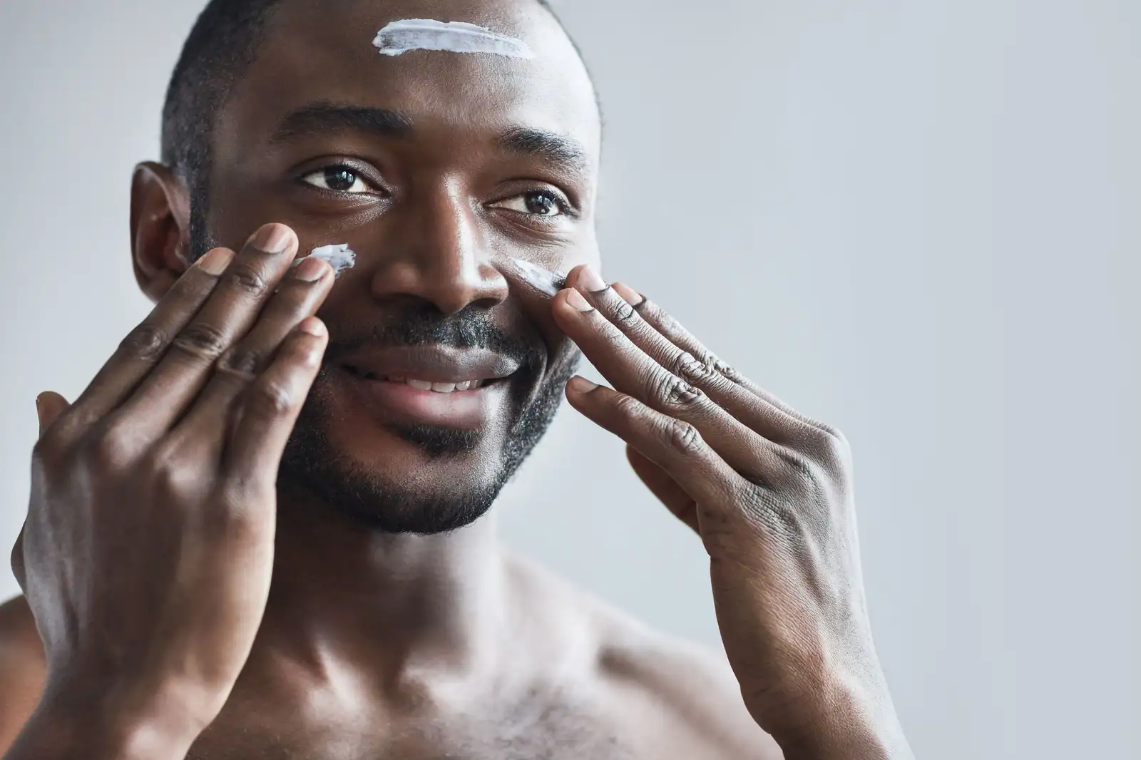 Jeune homme souriant appliquant une crème de soin sur son visage du bout des doigts