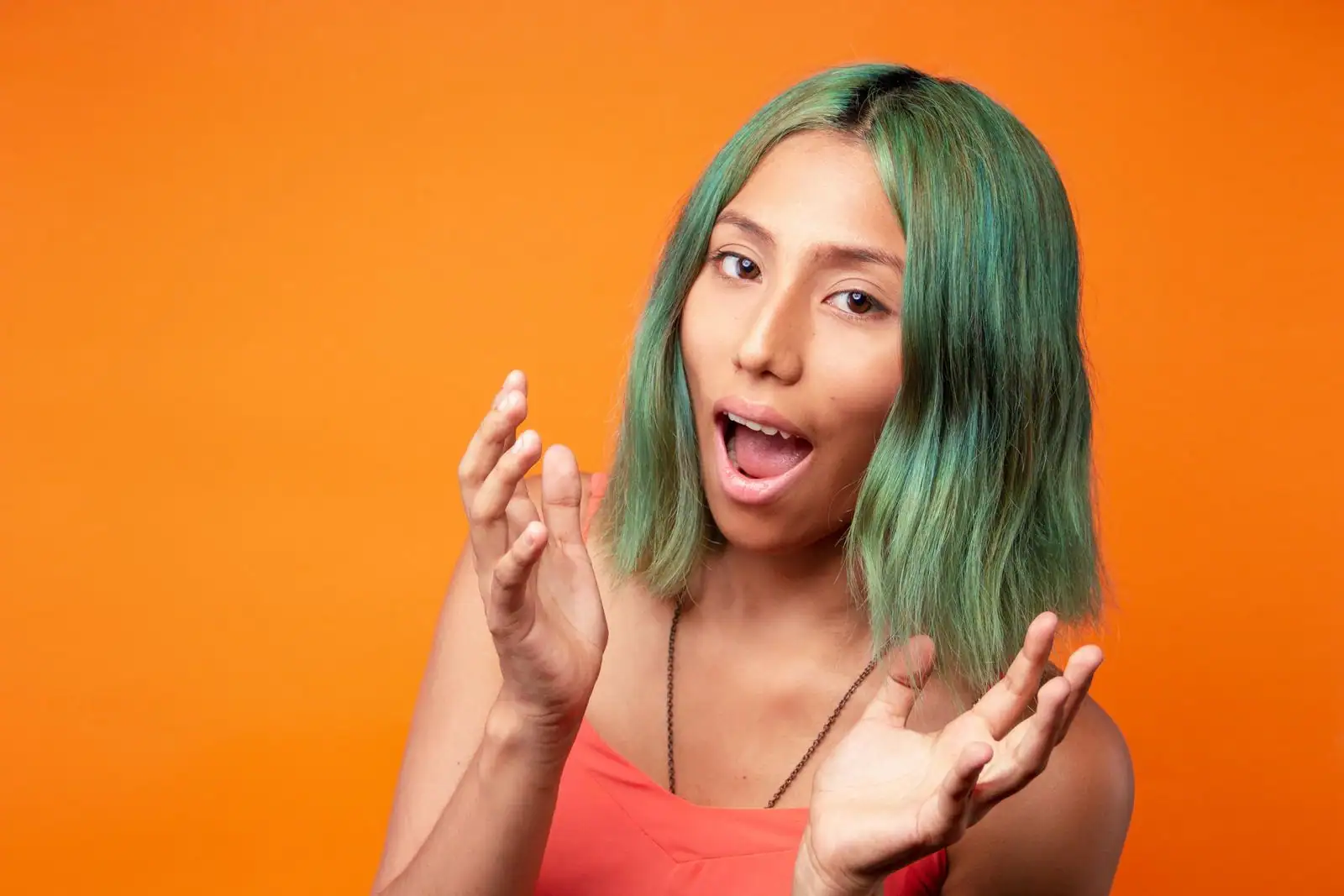 Jeune femme aux cheveux teints en vert, coupe carré mi-longue, posant sur un fond orange vif