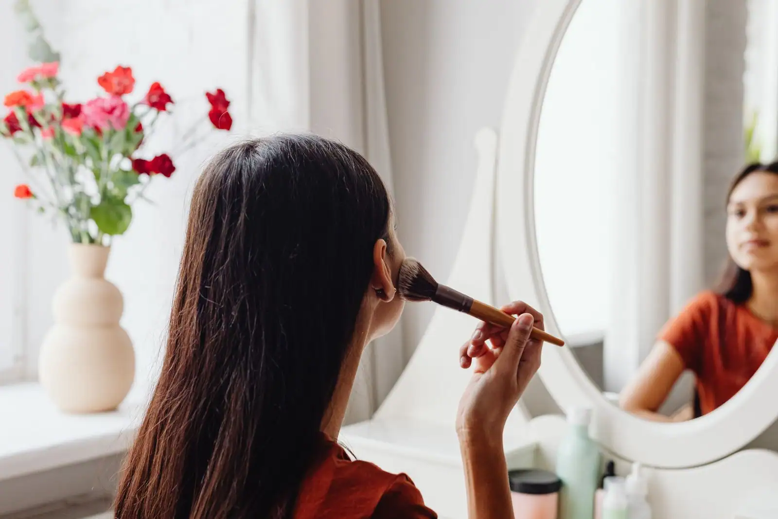 Femme se maquillant le visage au pinceau devant un miroir de coiffeuse, entourée de produits de beauté, dans une lumière claire