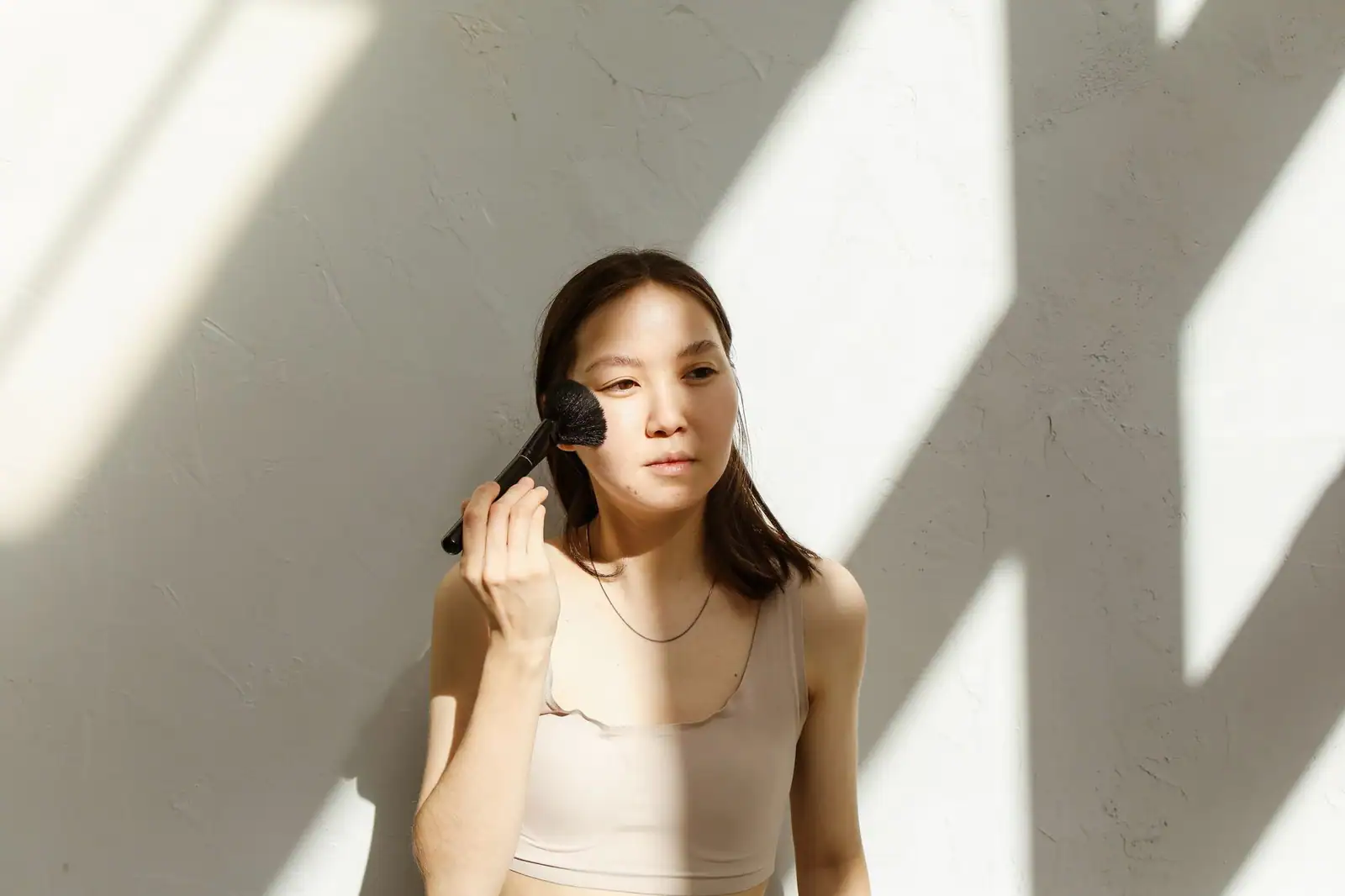 Femme asiatique appliquant du blush au pinceau sur la joue en pleine lumière, teint frais et naturel.