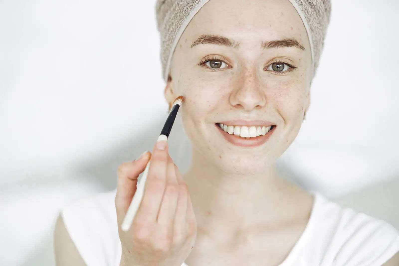 Jeune femme souriante au teint frais et naturel appliquant un peu de maquillage au pinceau, dans une lumière claire