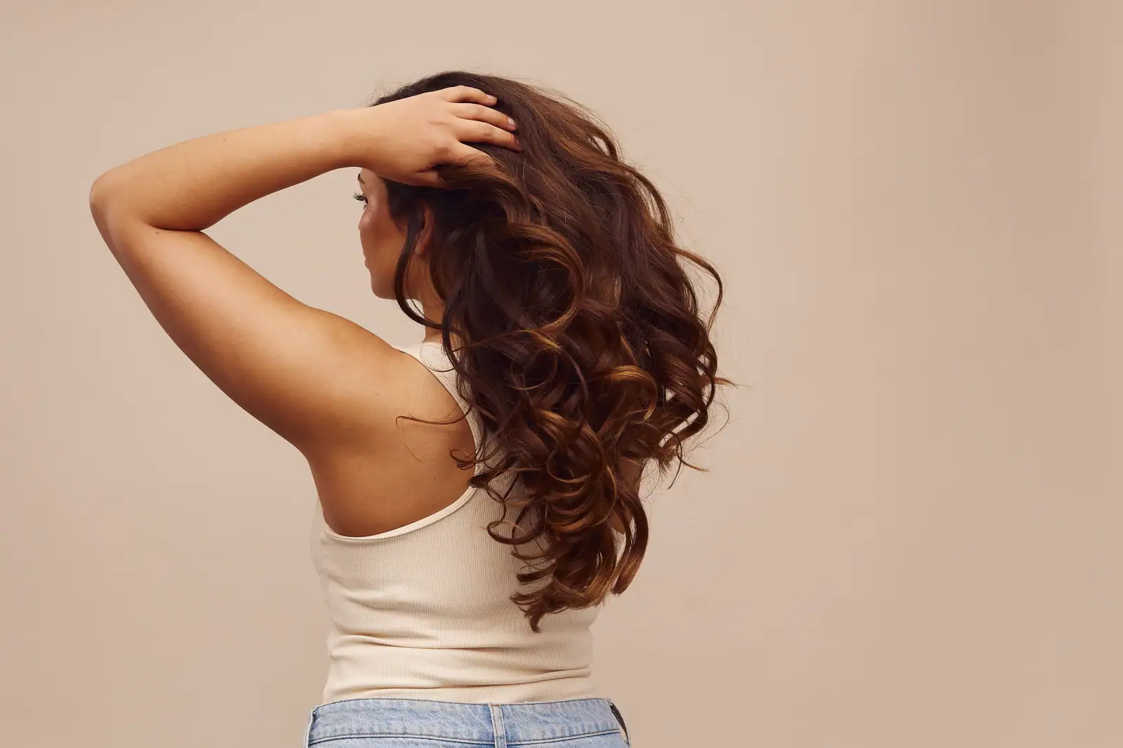 Femme de dos, longs cheveux bruns aux boucles bien dessinées, une main relevant la chevelure