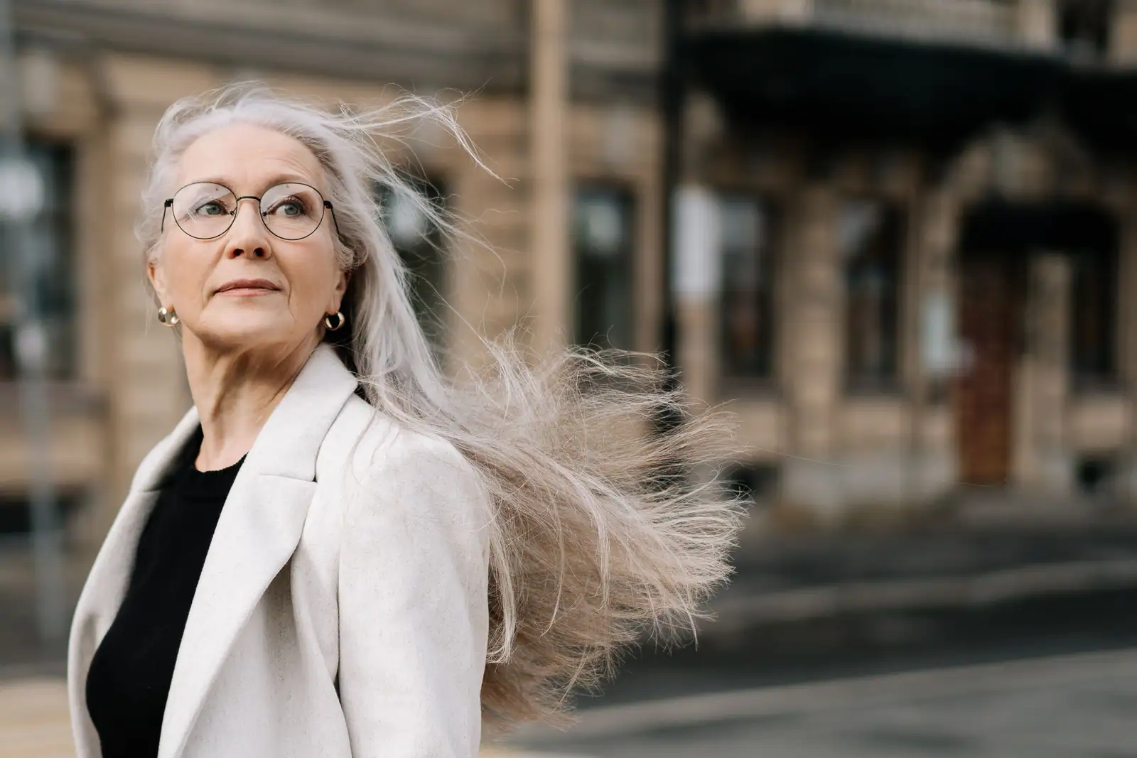 Femme aux longs cheveux gris argentés soulevés par le vent, lunettes rondes et blazer clair, en ville