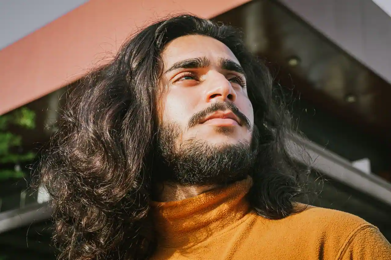 Homme aux cheveux bruns ondulés mi-longs retombant sur les épaules, photographié dehors en plein soleil