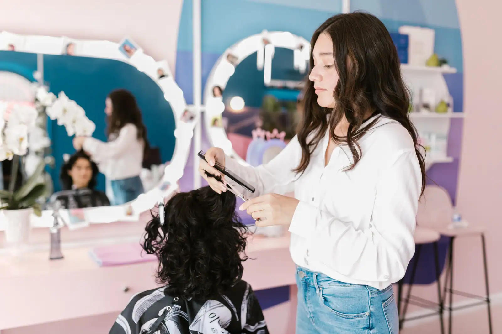 Coiffeuse aux cheveux longs coupant les cheveux bouclés d'une cliente dans un salon aux tons pastel