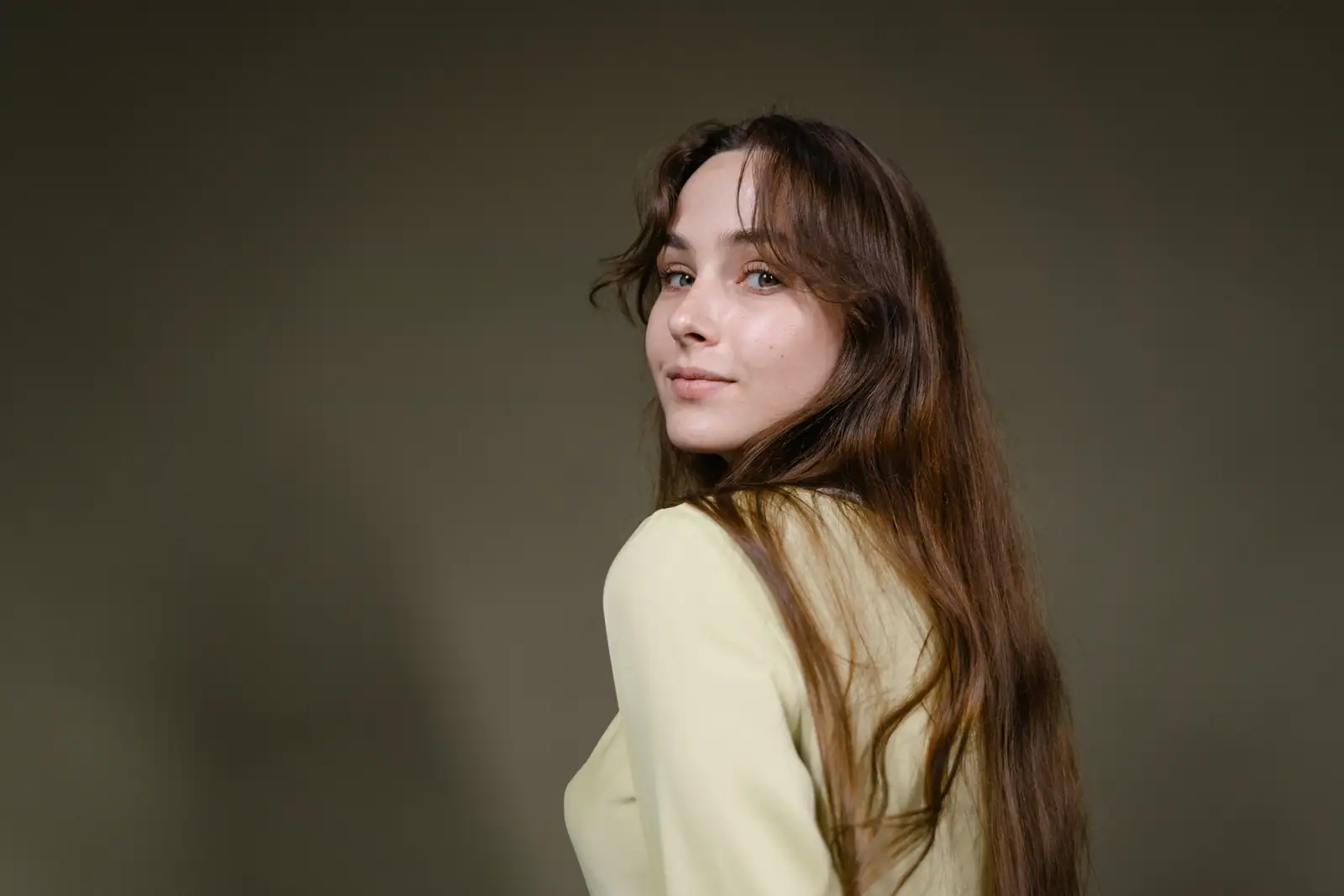 Jeune femme tournée sur l'épaule, longue chevelure brune dense retombant dans le dos, fond studio olive.