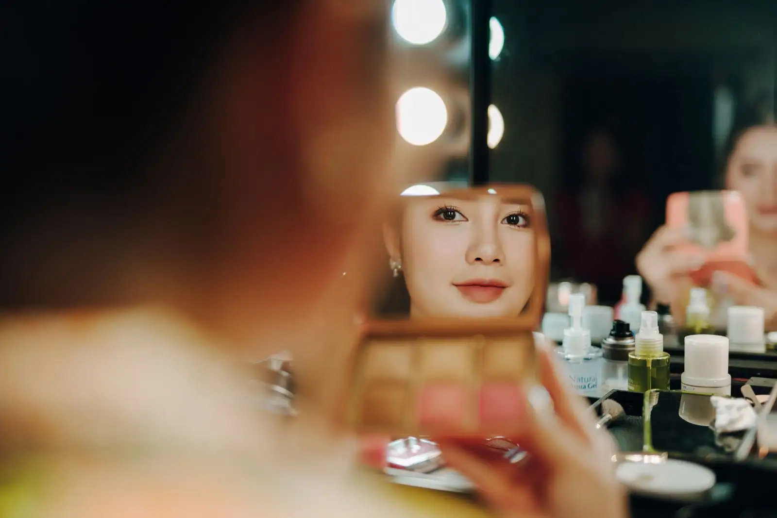 Jeune femme se maquillant devant le miroir éclairé d'une loge, une palette de blush rose à la main