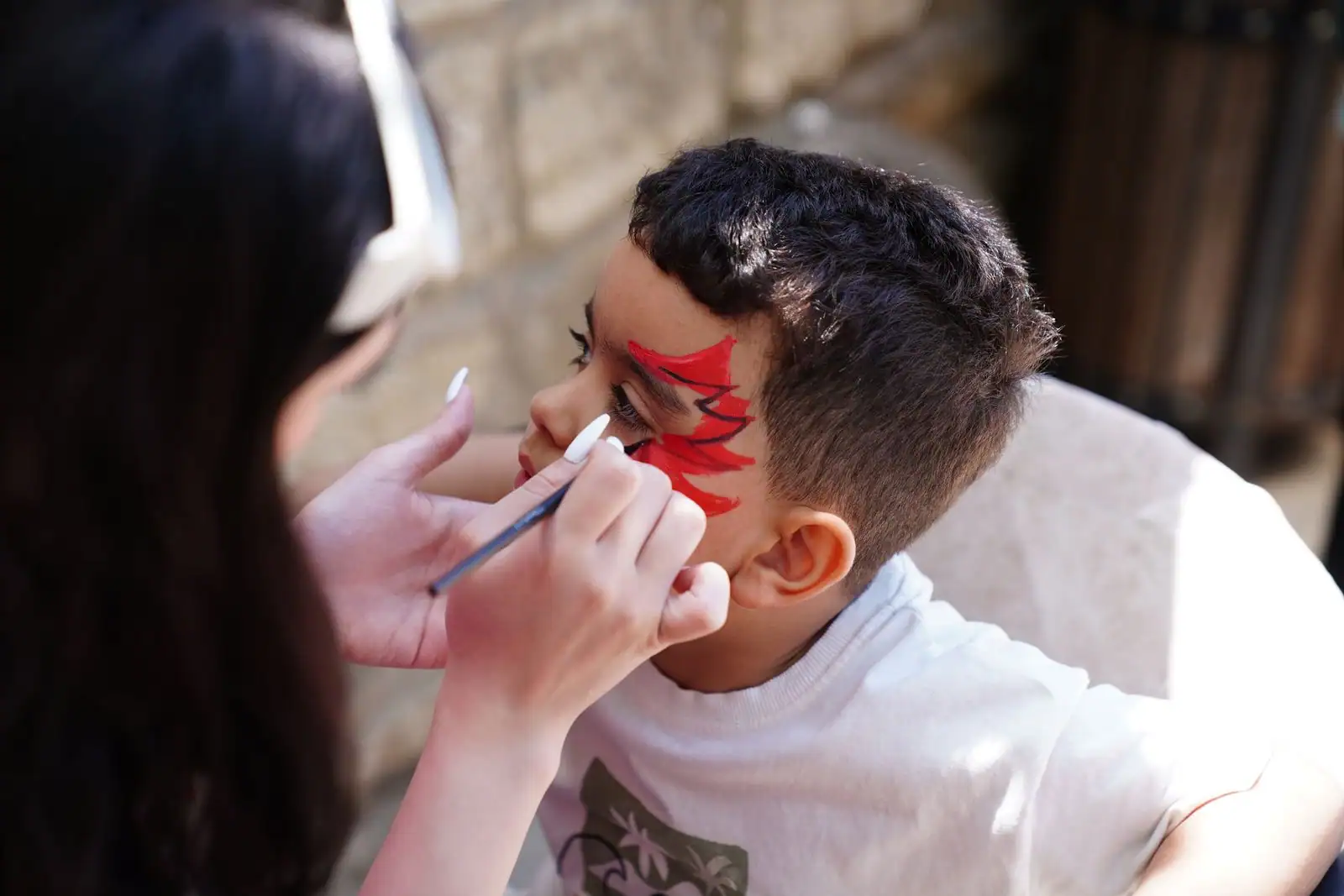 Maquilleuse traçant au pinceau fin un motif rouge cerné de noir sur le visage d'un jeune garçon