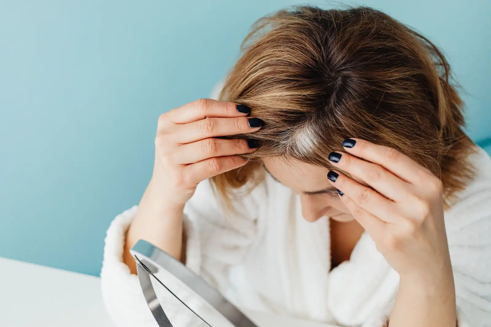 Femme écartant ses cheveux châtains devant un miroir, révélant une mèche blanche à la racine.