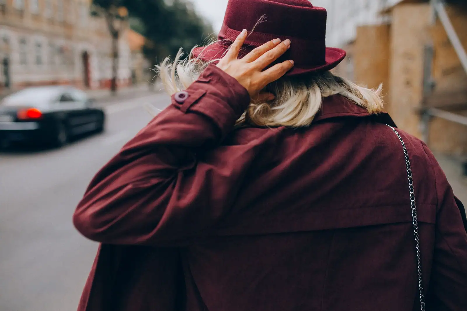 Femme de dos en manteau et chapeau bordeaux dans une rue urbaine, la teinte phare de la saison