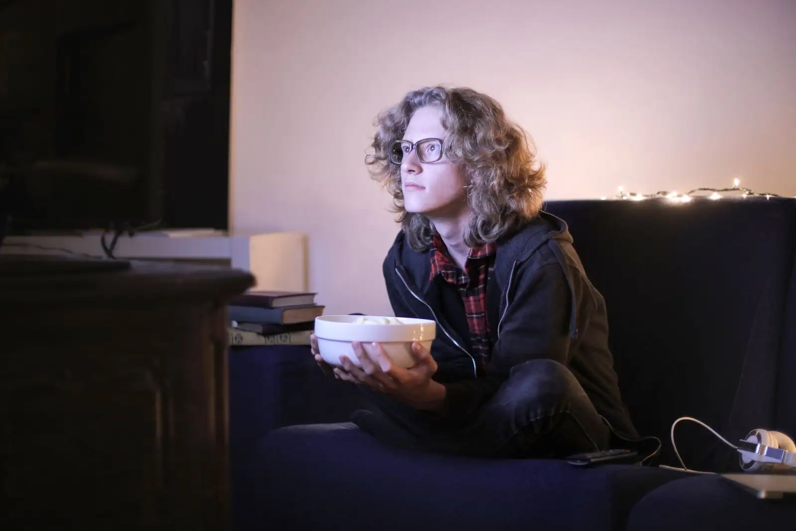 Personne installée en tailleur dans un canapé, un bol de pop-corn dans les mains, face à un écran en soirée